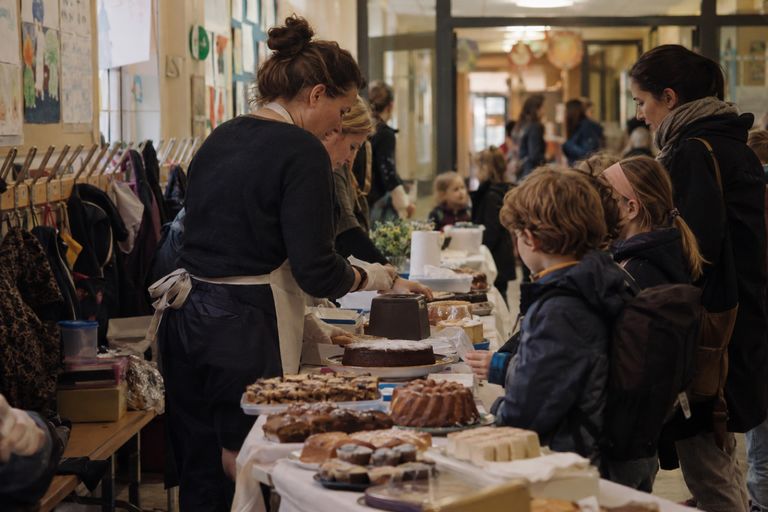 Kuchenverkauf im Schulflur: Zwei Helferinnen mit Schürzen verteilen Kuchenstücke an einem langen Tisch, davor warten Kinder mit Schulranzen und eine Mutter, an den Wänden hängen Kinderbilder.