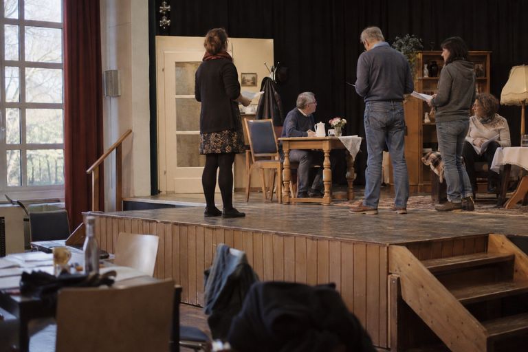 Theaterprobe auf der Bühne eines Vereinssaals: Fünf Laienspielerinnen und -spieler mit Textblättern stehen und sitzen in einem Wohnzimmer-Bühnenbild mit Tisch, Stehlampe und Schrank, vorn stehen Stühle mit abgelegten Jacken.
