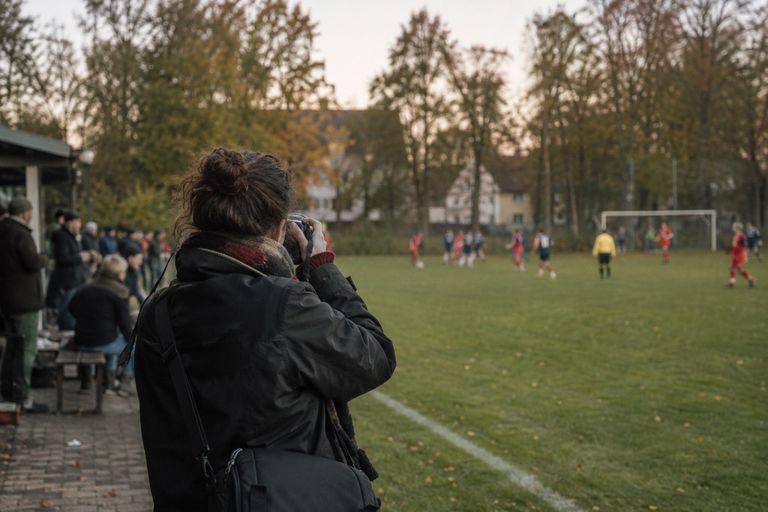 Eine Person mit Kamera fotografiert vom Spielfeldrand ein Fußballspiel, links schauen Zuschauerinnen und Zuschauer unter einem Vordach zu.